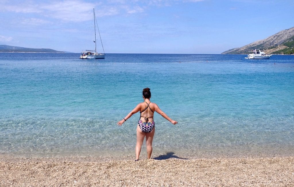 Kate in a one-piece bathing suit, shot from behind, alone on a pebble beach in Croatia, the ocean looking clear and aqua. She holds her hands out and walks into the sea.