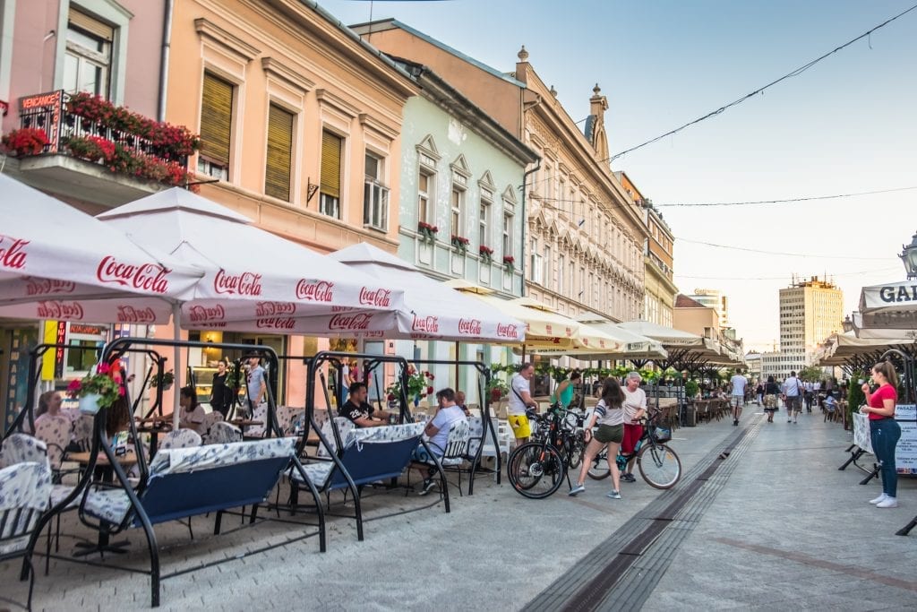 Street cafes in Novi Sad. The buildings in the background are pastel shades of pink, orange, and mint green.