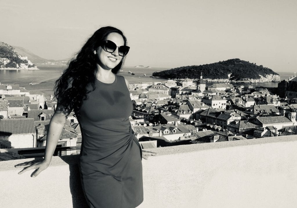 Kate stands in front of the Old City of Dubrovnik, rising behind her in terra cotta roofs and church towers, green wooded Lokrum Island in the distance.