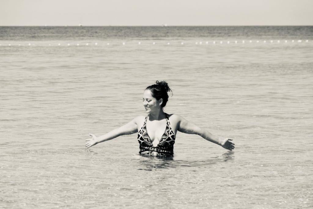 Kate stands in waist-deep water with her hands splayed, smiling and looking to the side, in Croatia.