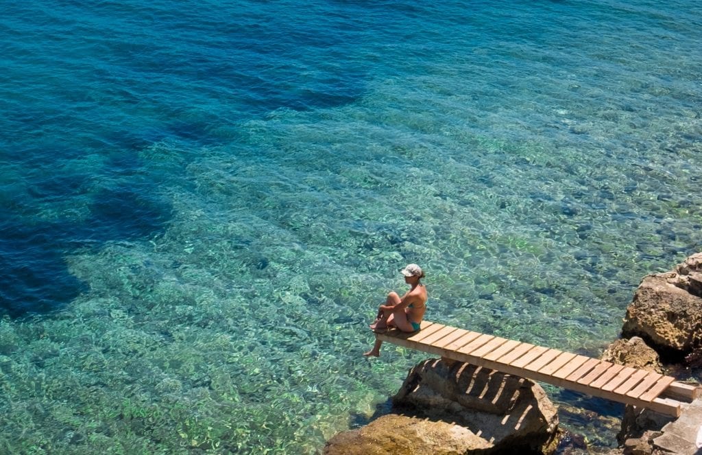 A woman sitting on a plank jutting out into the clear blue-green water of the sea.