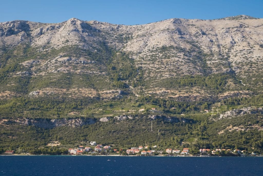The view of Pelješac from Korčula town: a zoomed-in view of a tiny small town of white homes with orange roofs, and straight upward is a mountain, lots of greenery near the bottom, and more and more empty gray stone near the top.