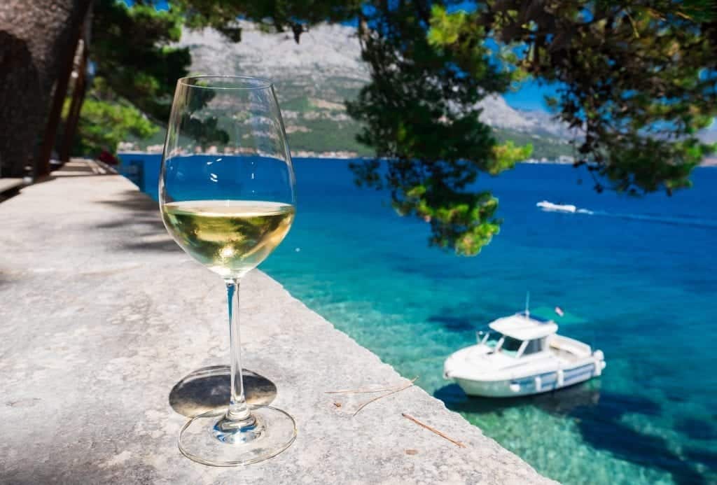 A glass of golden Grk wine in the foreground on the edge; in the background, bright blue-green water and a small boat.
