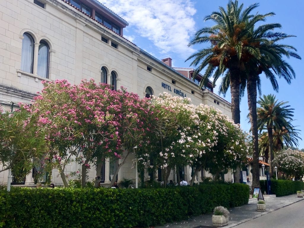 The former Hotel Korcula de la Ville, a white stone building with several small trees in front, blooming in pink and white.