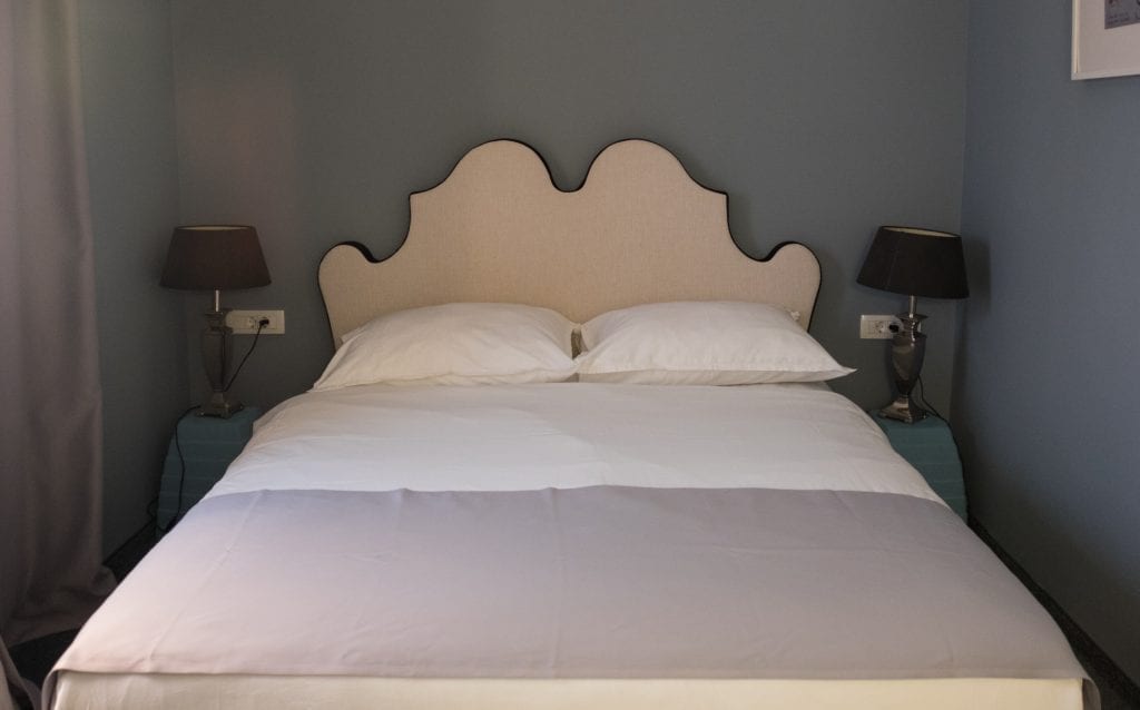 A white bed with a curvy white fabric headboard in the hotel, small tables with black lamps on each side.