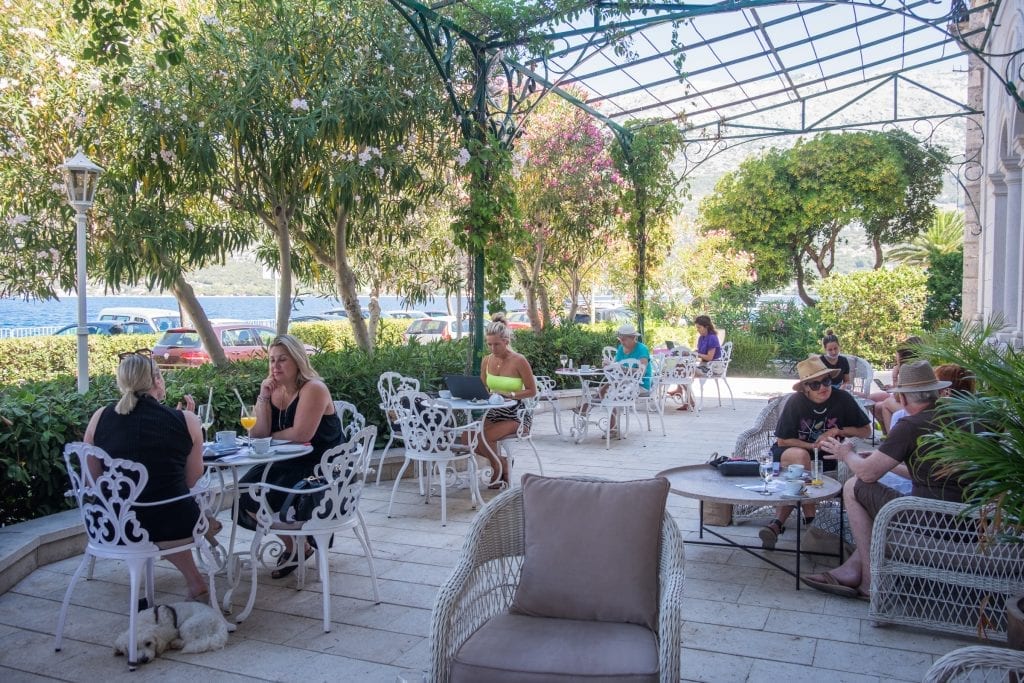 An outdoor terrace filled with several white wrought-iron tables, some of them with people sitting at them enjoying drinks or working on laptops.
