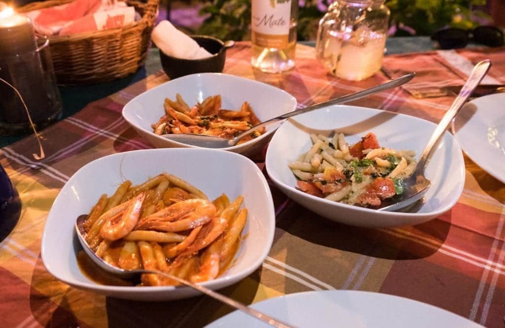 Three plates of oblong hand-rolled pasta: one with shrimp, one in tomato sauce, one in almond pesto with cherry tomatoes.