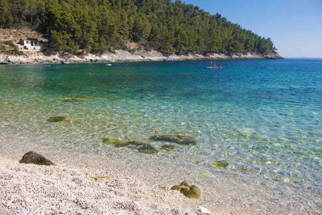 A pebbly beach with clear blue-green water, a few people on a stand up paddle board in the distance.