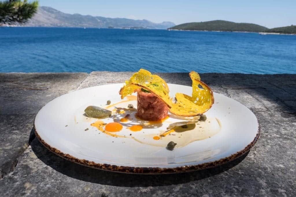 A fancy plate of food at LD restaurant on the edge of the old town overlooking the blue ocean and islands in the background. There's a Vitello tonnato looking like a beef tartare, a curved thin piece of stiff bread curved around the top, with cured caper leaves and drops of cured egg yolk on the plate.