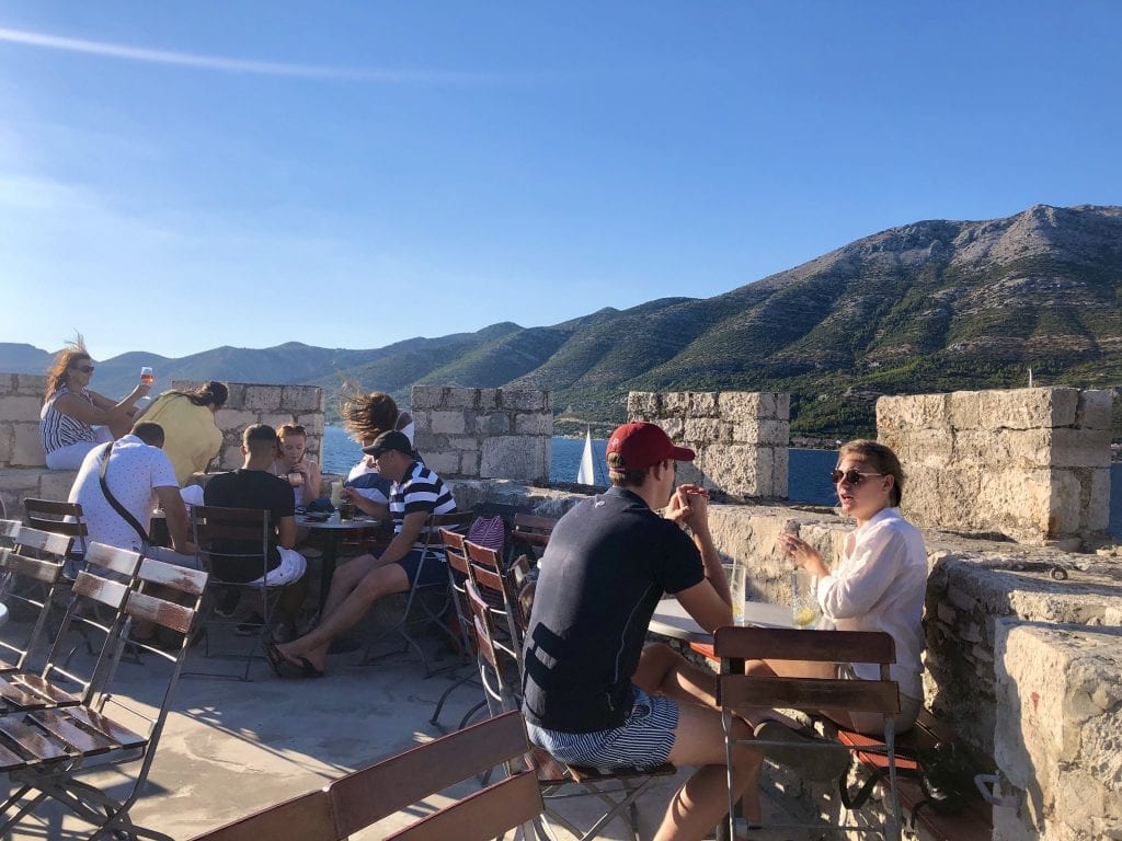 People sitting outside in Massimo Bar on Korcula, sitting at distanced tables in what looks like the top of a castle, overlooking the mountains on shore.