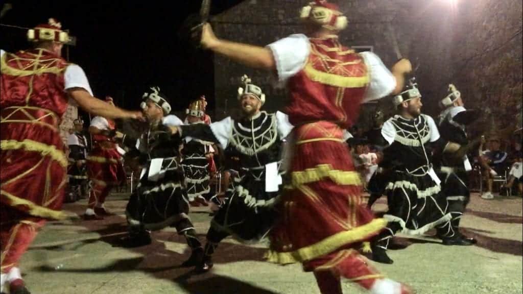 Several men in red and black medieval costumes doing a sword dance in circles around each other.