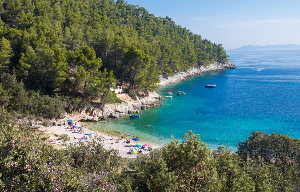 A small white pebble beach leading into the bright blue ocean in Korčula, Croatia. It's a narrow inlet surrounded by rocky coastline topped with bright green trees.