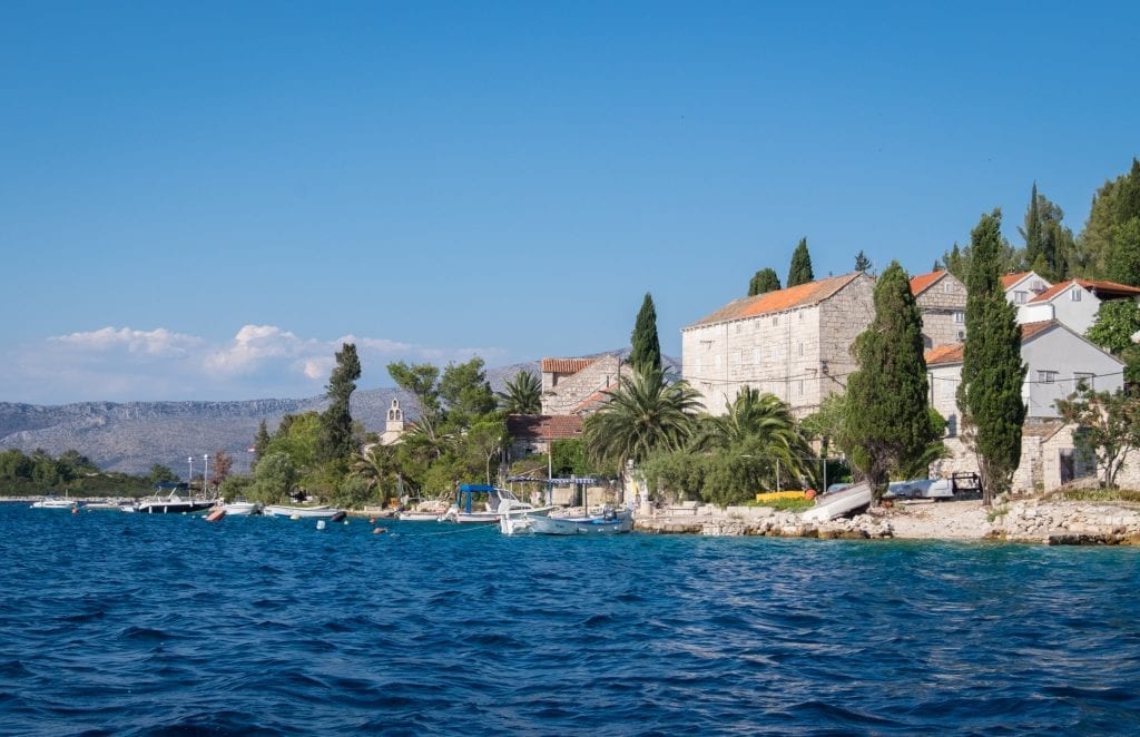 A close-up of Vrnik island with boats anchored to shore, stone buildings with terra cotta roofs, and lots of palm and cypress trees.