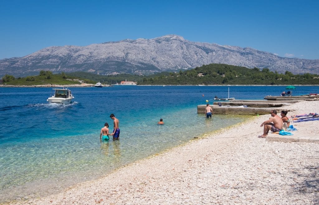 A quiet beach with a father helping his son into the blue-green water, a speedboat and mountains in the background.