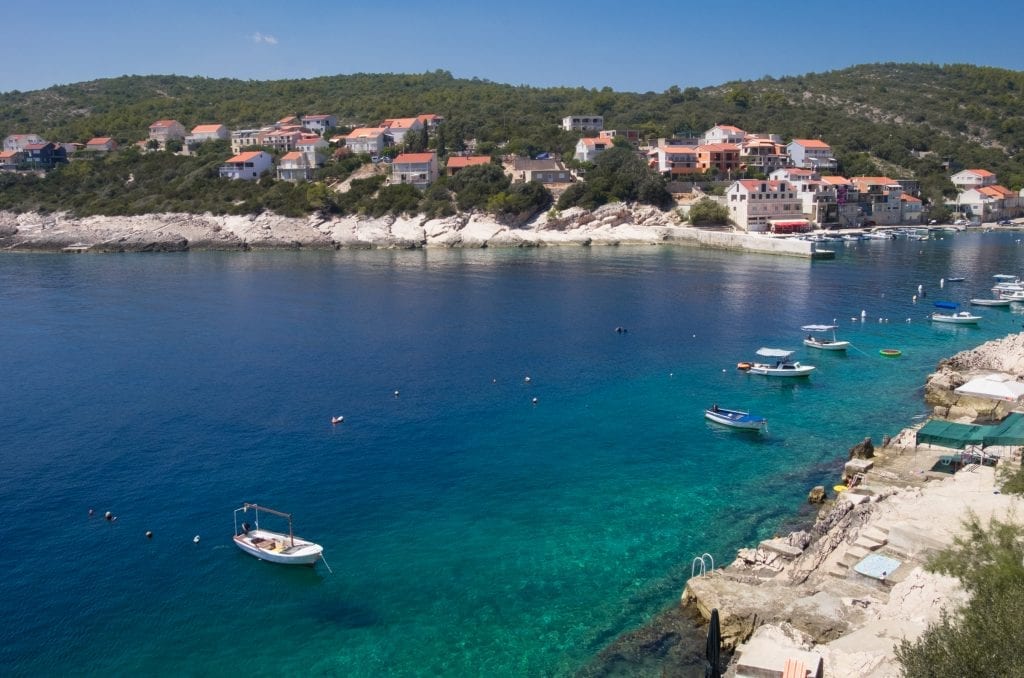 Zavalatica: A view from above a bay in shades ranging from navy to teal, filled with small speedboats, edged with a rocky waterfront, white houses with orange roofs further back from the coastline.