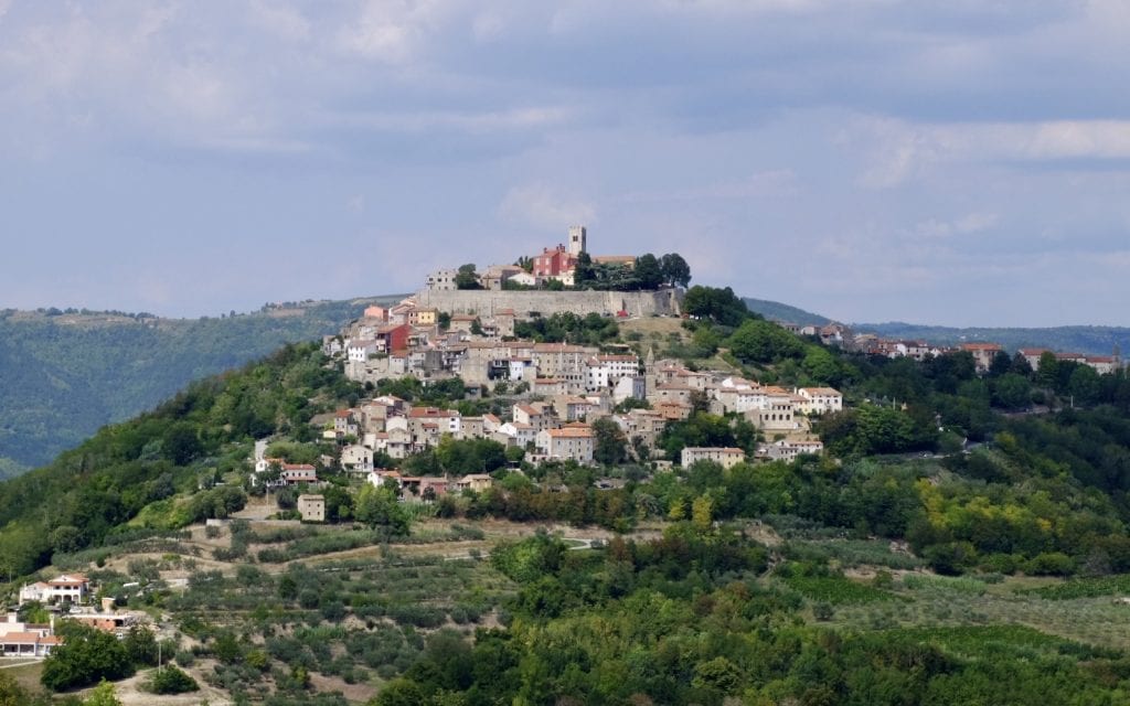 Motovun: a hill town perched on top of a bright green hill. A small wall and several rows of beige, white, and brown houses on top.