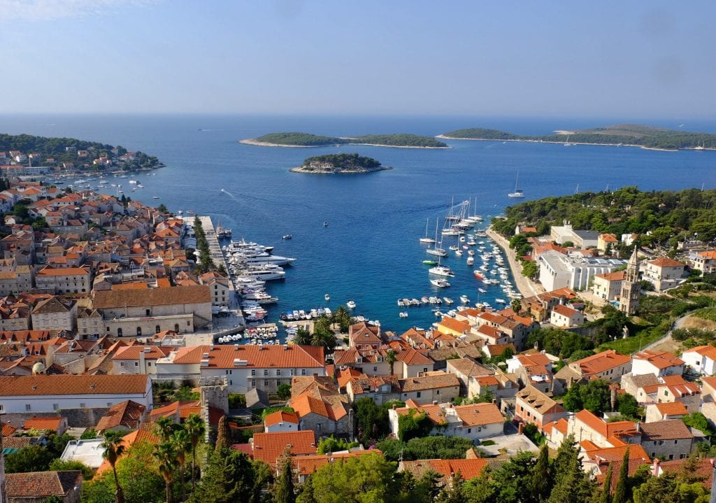 The view of Hvar from the Spanish Fortress: orange-roofed buildings and green trees surrounding a three-sided harbor edged with white boats of all sizes, the Pakleni islands in the distance, shaped like green droplets in the sea.