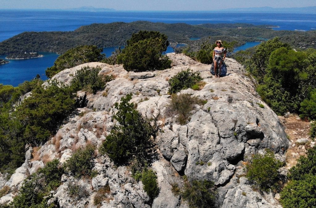 Kate standing on top of a rocky formation in Mljet, views of the island in the distance, shot from a drone above.