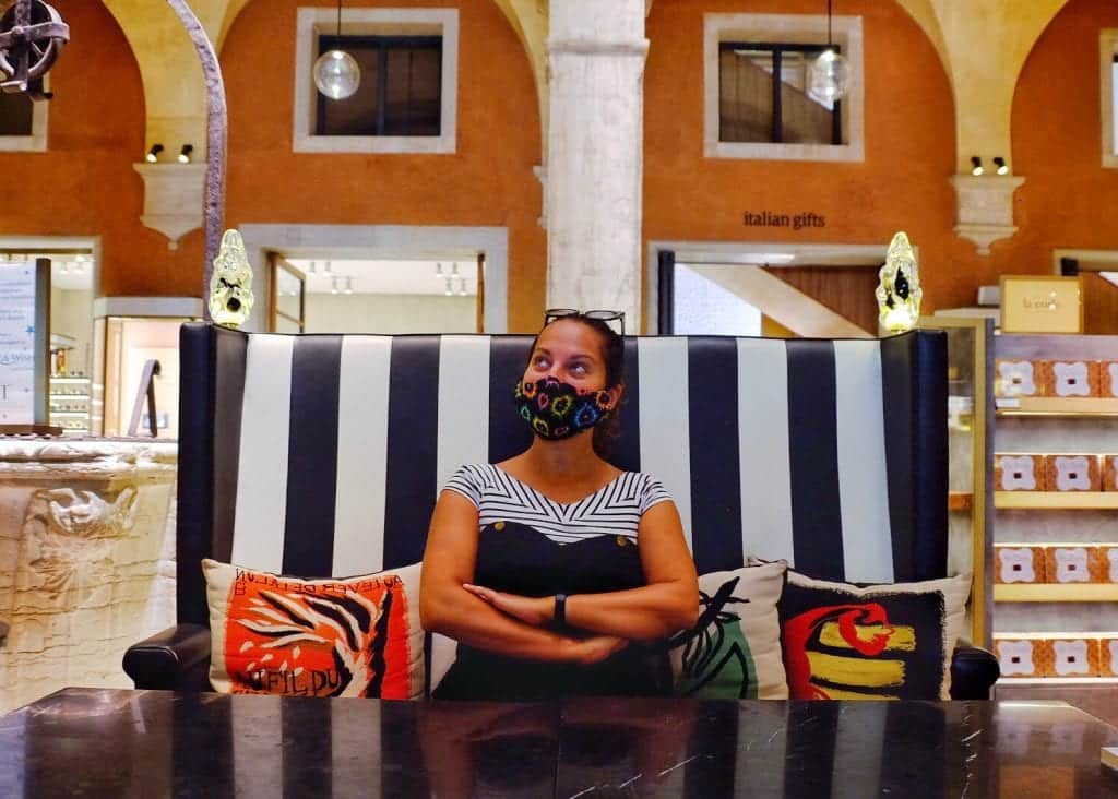 Kate sits on a black and white striped booth in a department store in Venice, columns behind her. She has her arms crossed and she's wearing a black face mask covered with splashes of color, and she's looking upward.