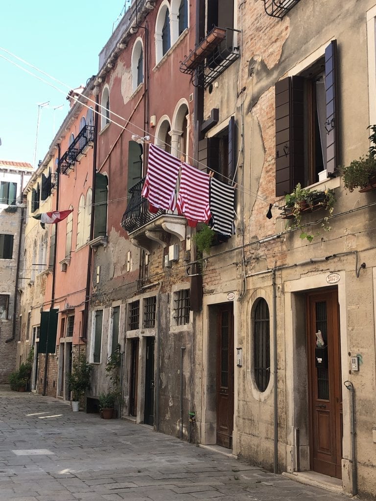 A street in Venice lined with homes. Out one window hangs laundry: two red and white striped shirts and a black and white striped shirt. The uniform of a gondolier.