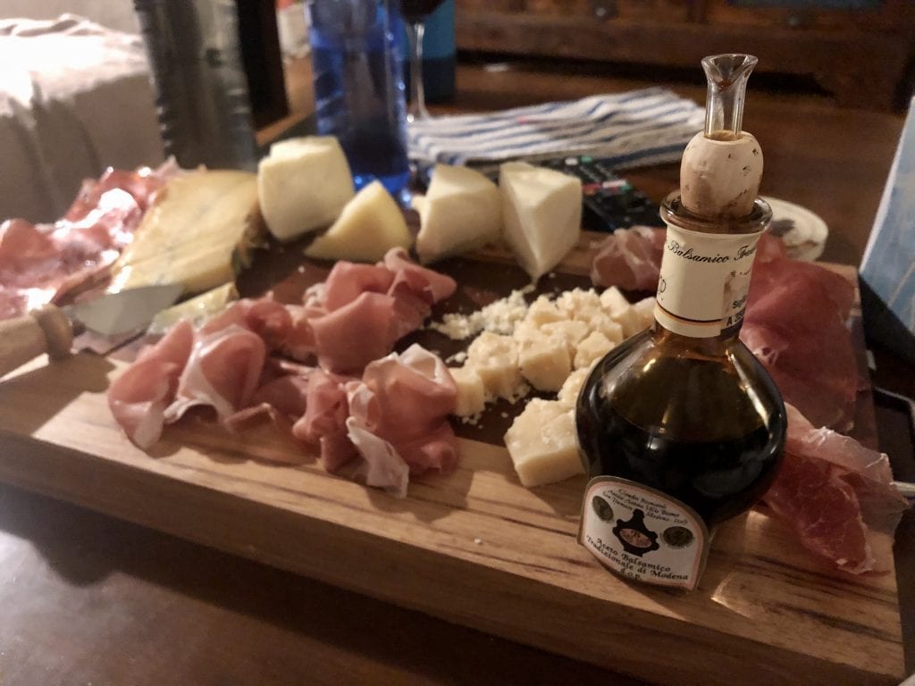 A wooden cutting board topped with various cured meats, cheeses, and a bottle of traditional balsamic vinegar from Modena.