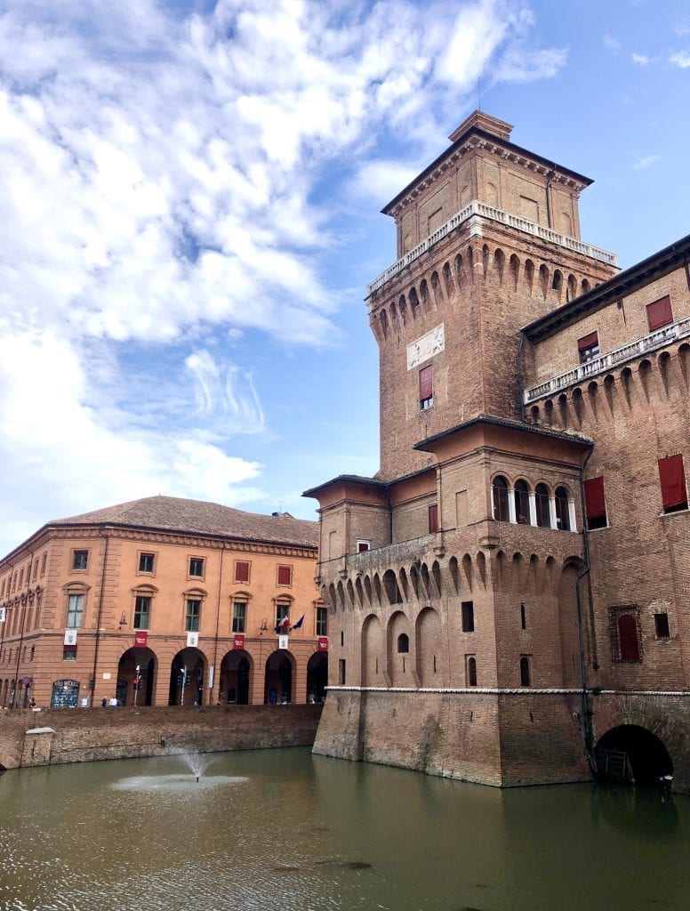 Ferrara's red brick boxy castle, set in the middle of a green moat, underneath a blue partly cloudy sky, an orange building with porticoes in the background.