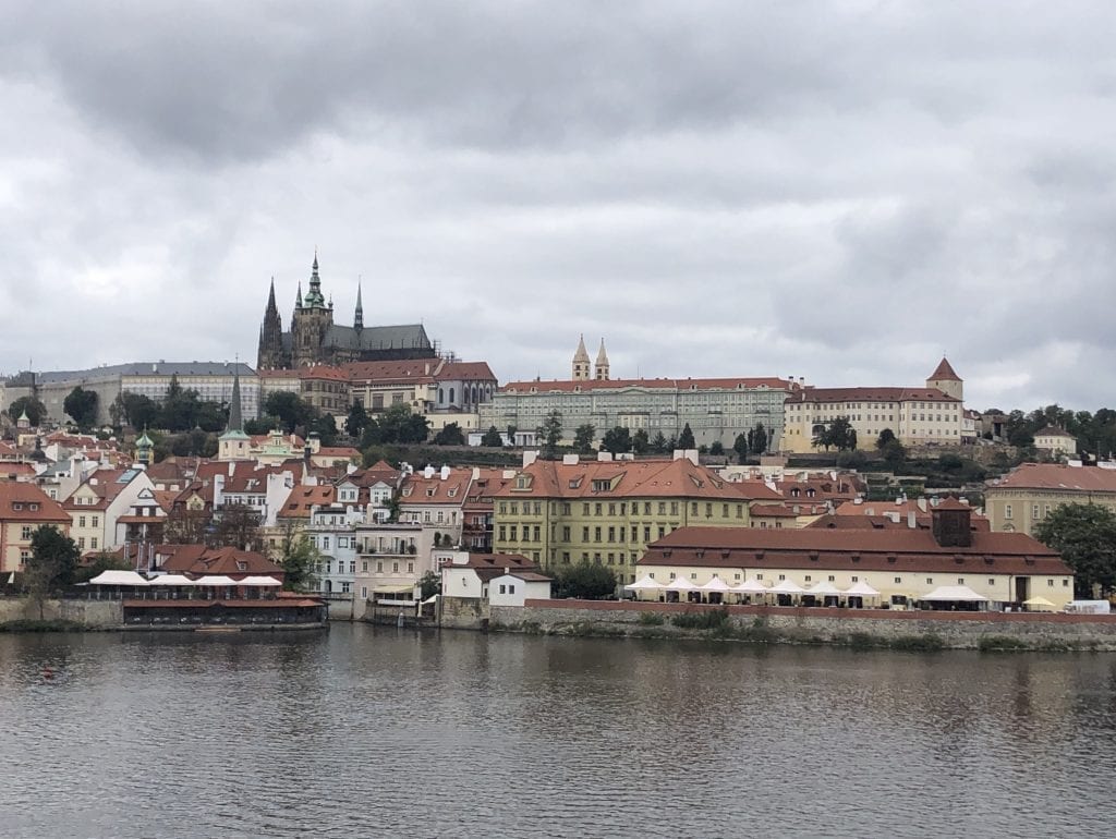 A view of Prague Castle from the Charles Bridge on a cloudy day, the spires sticking straight up from the cathedral, set on a dark gray river.