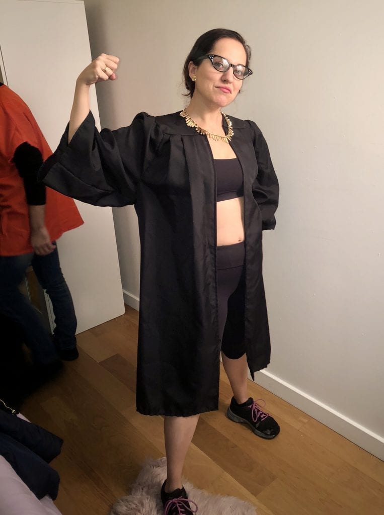 Kate's 2018 Halloween costume: Ruth Bader Ginsberg at the gym! Kate wears black glasses, an open black robe, a bright gold collar-like necklace, a black sports bra, black yoga pants, and black sneakers. She holds her arm up to show off her muscle.