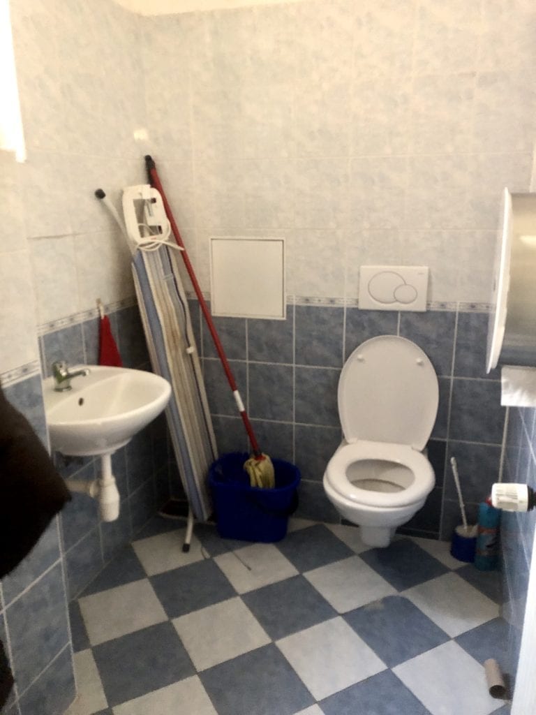 A tiny bathroom with toilet, sink, and an ironing board and mop leaning against the wall.