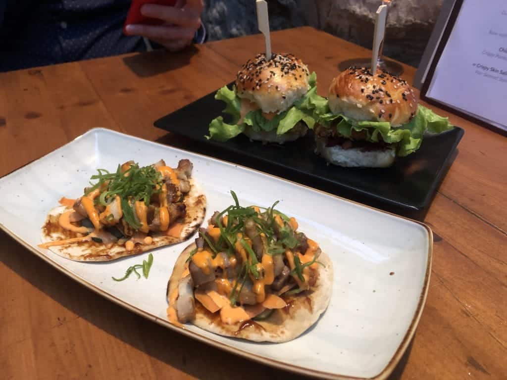 Two mini pork belly tacos on a white plate and two mini Korean fried chicken burgers on a black plate all on a wooden table at Azur  restaurant in Dubrovnik