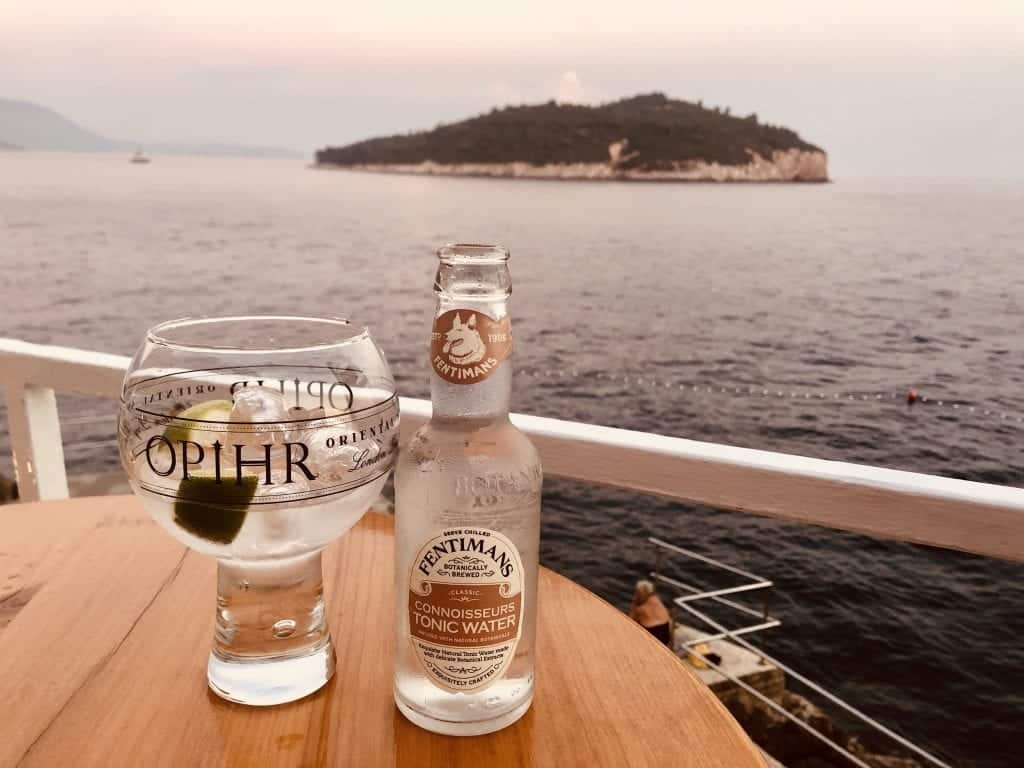 A large glass and a bottle of tonic water are placed on a wooden table top with the sea and an island in the distance behind them