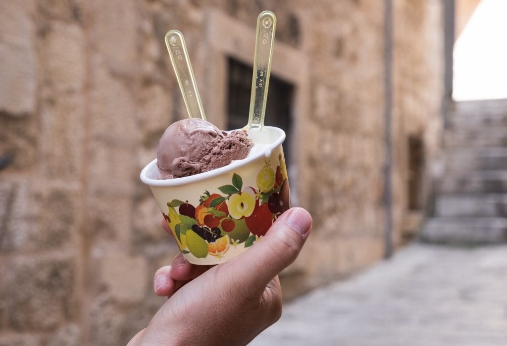 A hand holds a cup of gelato with two spoons against a blurry backdrop of Dubrovnik's Old City