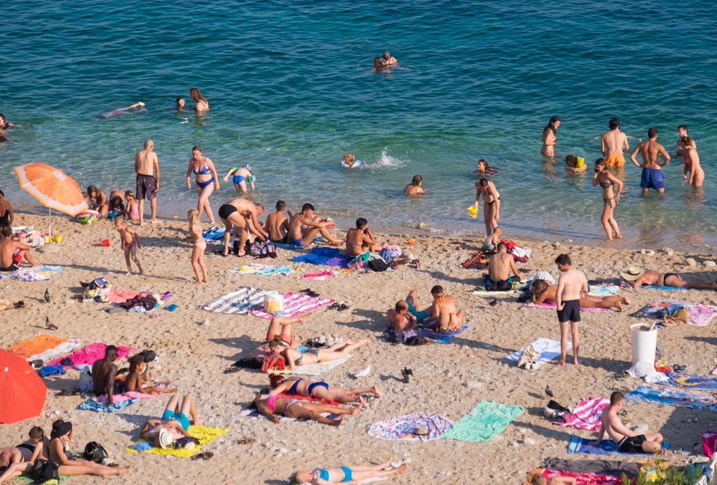 Lots of people on the beach, walking around, lying on beach towels or wading into the water.