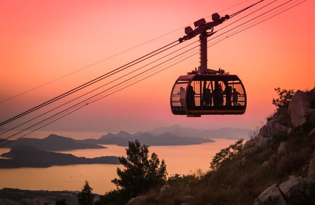 A cable car ascending a mountain, lit up from behind in a pink sunset. You can see islands in the distance.