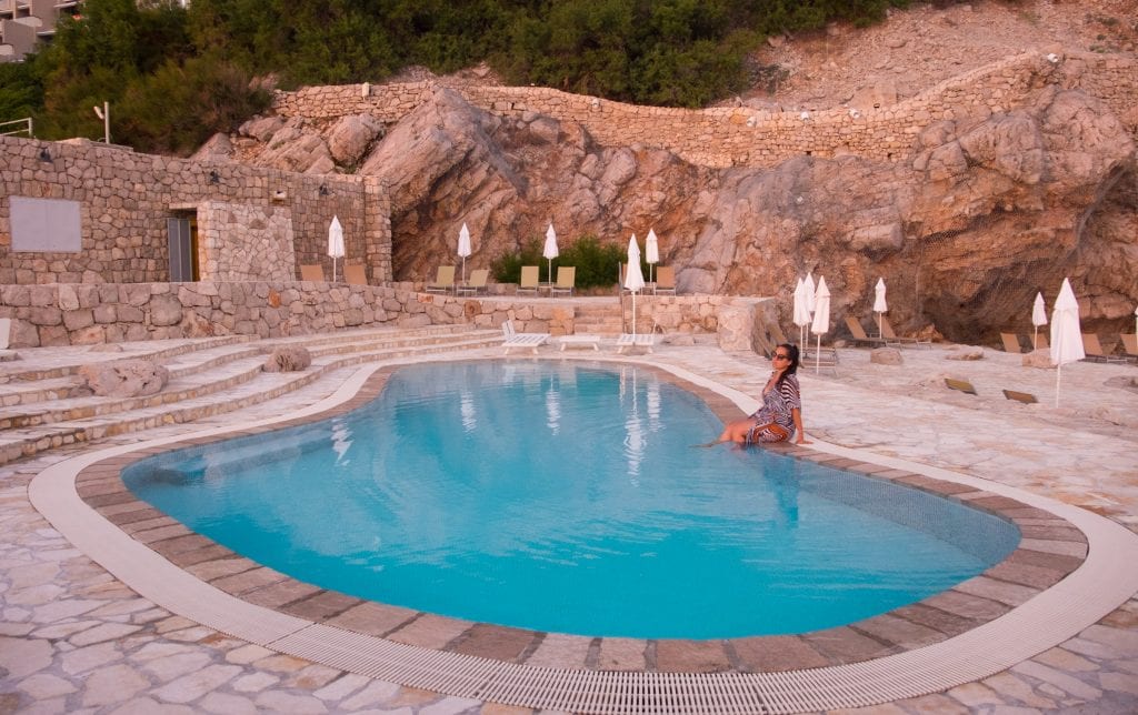 Kate sitting on the edge of the pool at Hotel Dubrovnik Palace surrounded by sandy rocks with umbrellas and beach chairs in the background