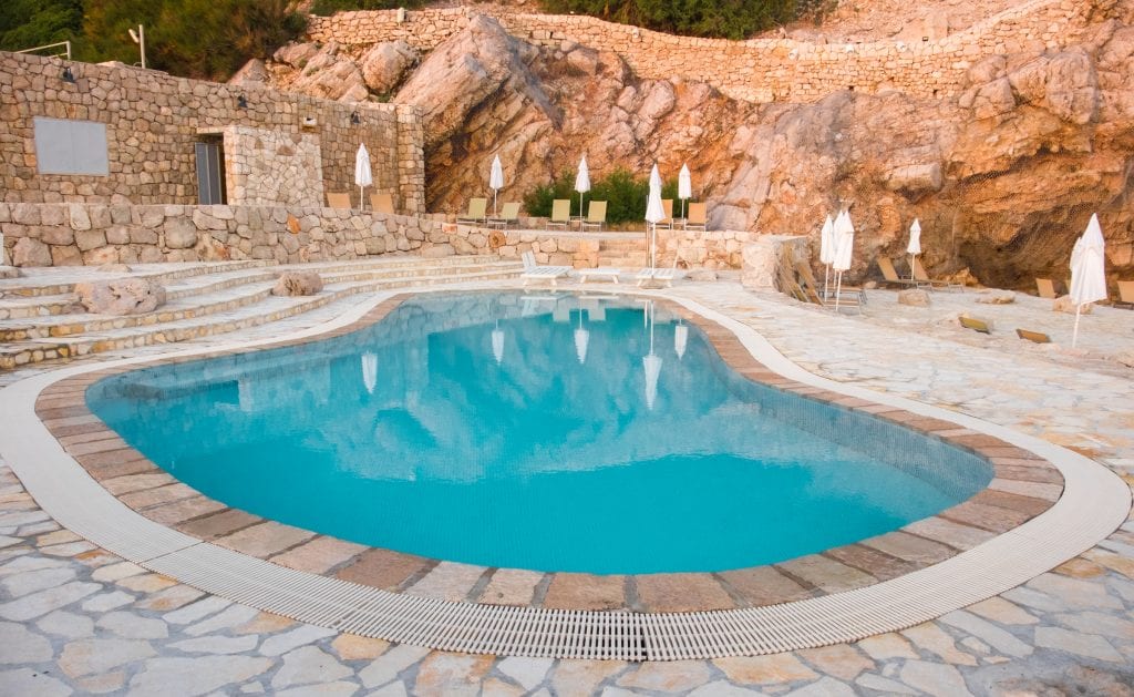A bright blue pool surrounded by rocks and stones in Dubrovnik, Croatia. Lounge chairs and white umbrellas are dotted around the perimeter.