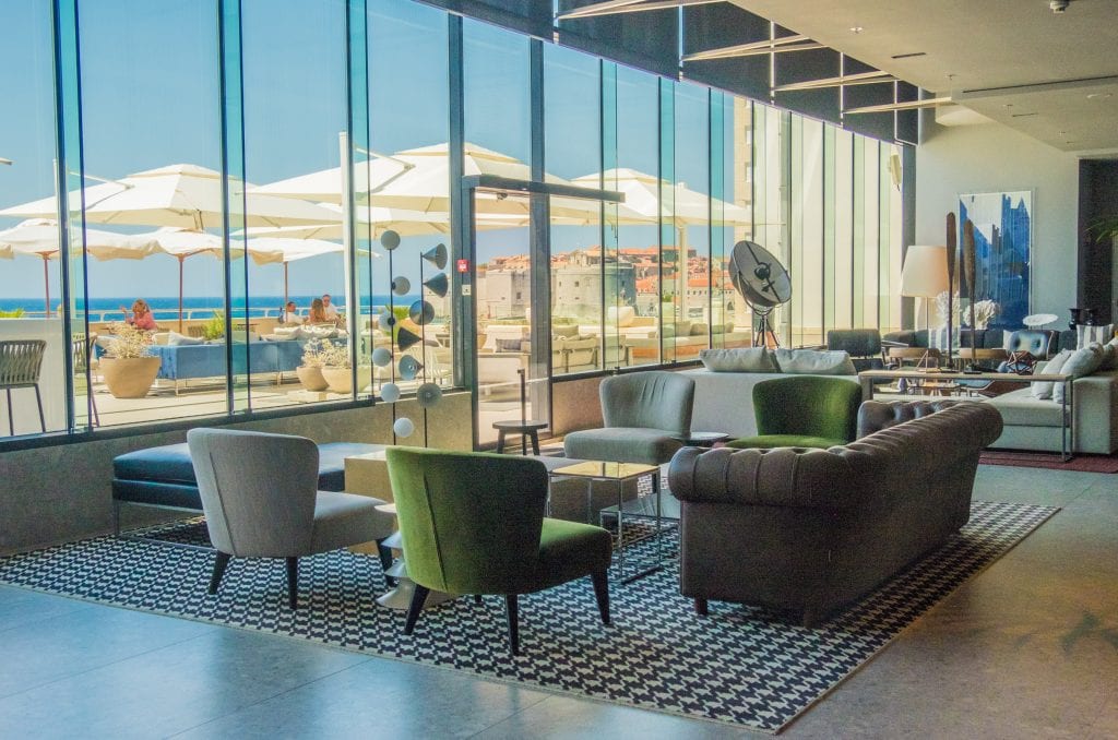 Lounge inside Hotel Excelsior with lots of comfortable chairs and couches, and huge floor to ceiling windows that look out over the patio and Adriatic Sea