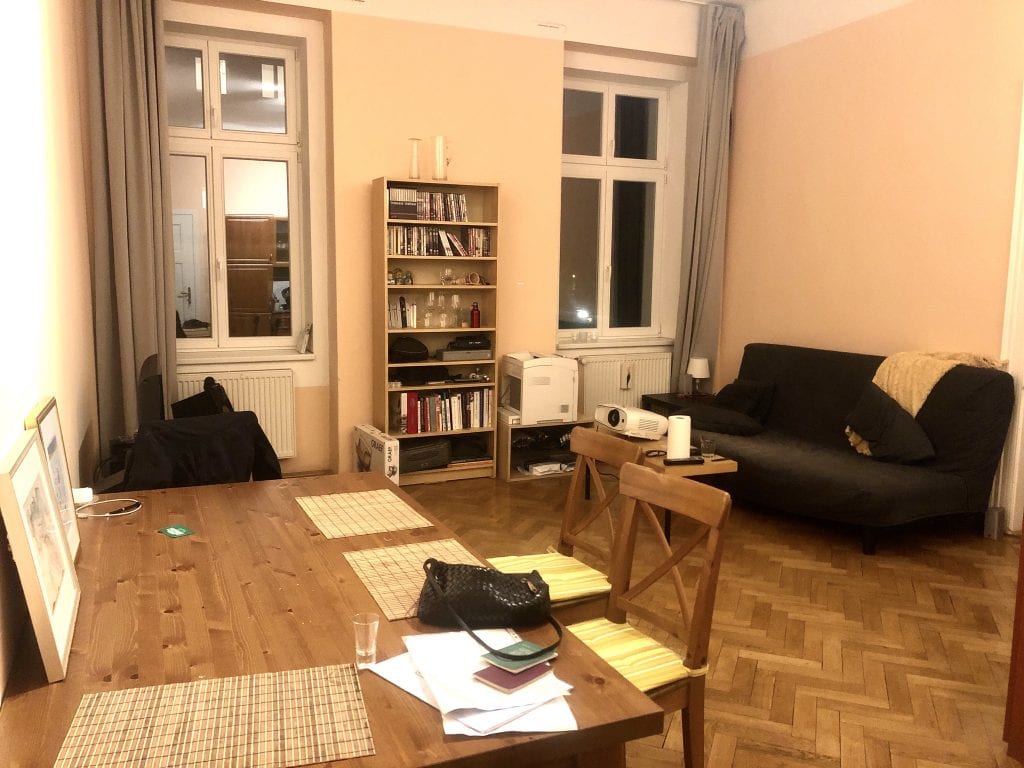 The living room: a big wooden dining table in the foreground, a bookcase with wine glasses, books, and random items in the background, with a gray futon and plastic-wood IKEA table with a projector on it.