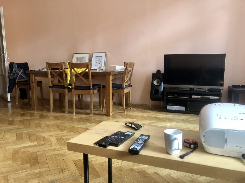 View across a living room: in the foreground, a cheap wood-and-plastic IKEA table; in the background, a large wooden dining table and a TV on a black TV cabinet.