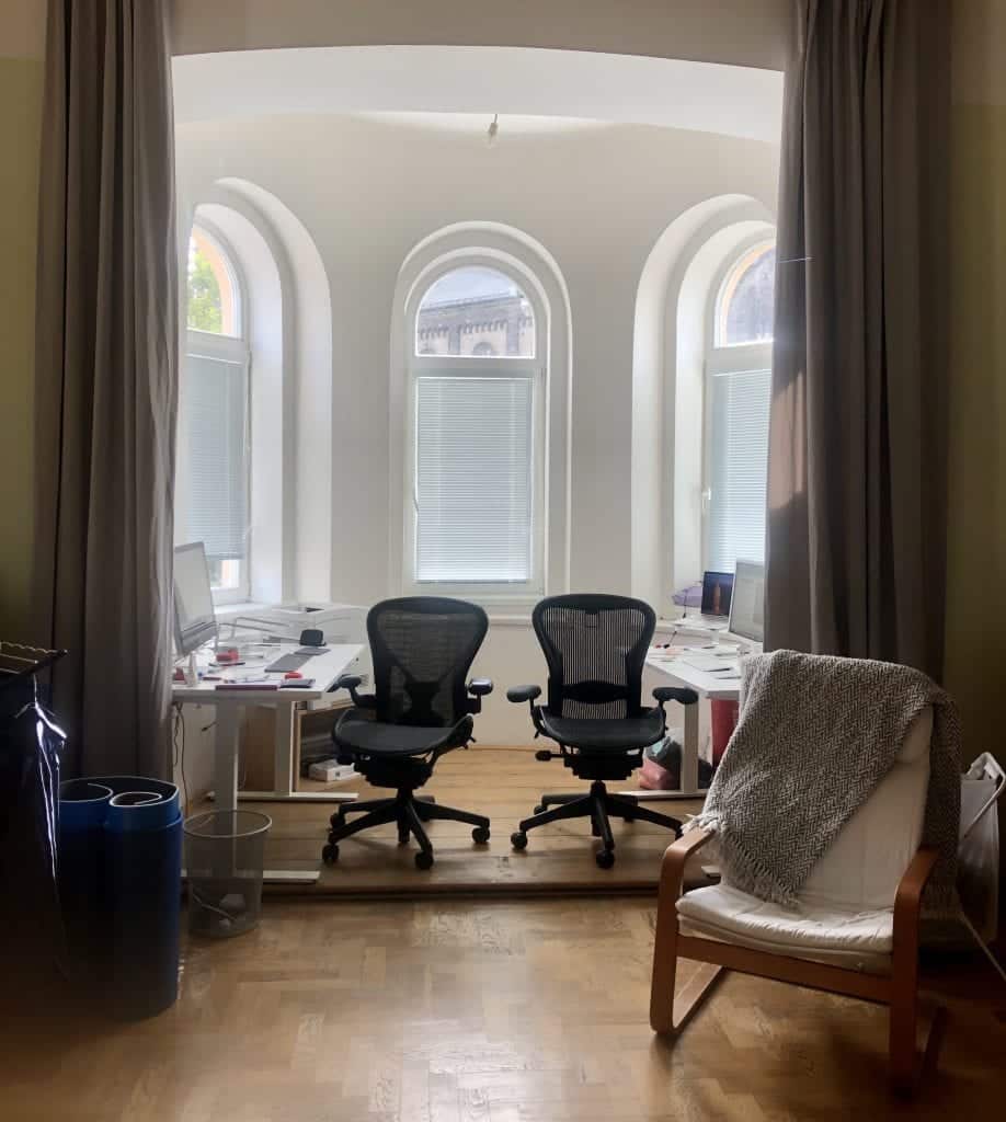 A small white room with three windows curved on top. It's a round room -- a turret! Each side has a white desk with monitors and computers on it, as well as a black office chair. Long gray curtains on each side.