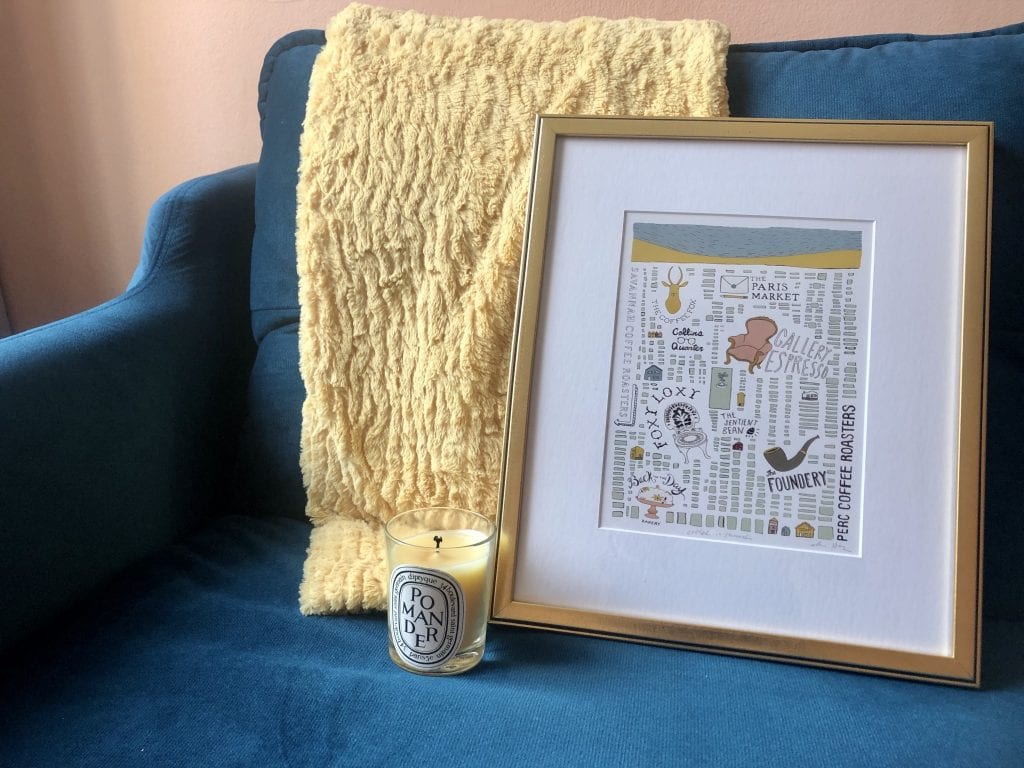 A gold-framed map of coffeeshops in Savannah, as well as a Pomander Diptique candle and a gold blanket on top of the teal couch.