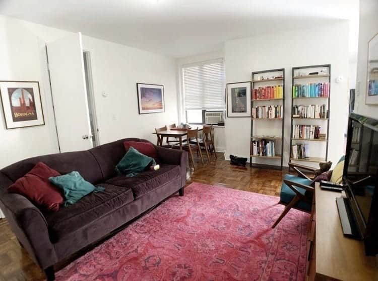 Kate's New York Living Room: a bright pink Persian rug, dark purple couch with maroon and teal cushions, and bookshelves in the background with a rainbow of books on top.
