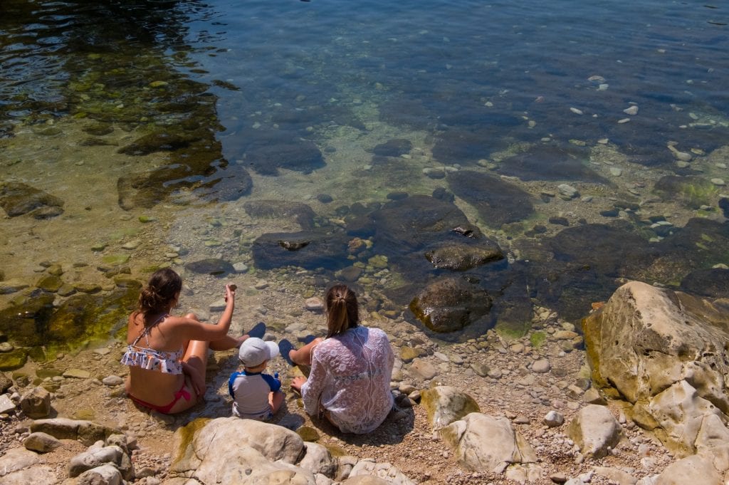 Two women and a small child sit on rocks in the ocean off of Lokrum Island in Croatia
