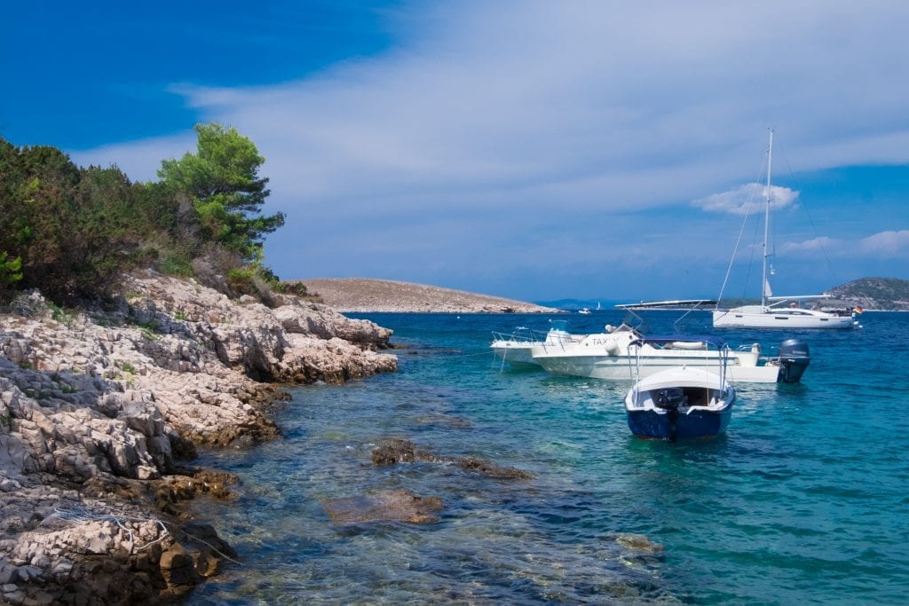 The rocky coastline of the Pakleni Islands, the water bright turquoise, three sailboats moored just off shore.