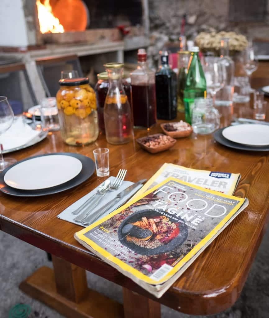A dining table set with drinks, glasses, plates, cutlery and two travel magazines