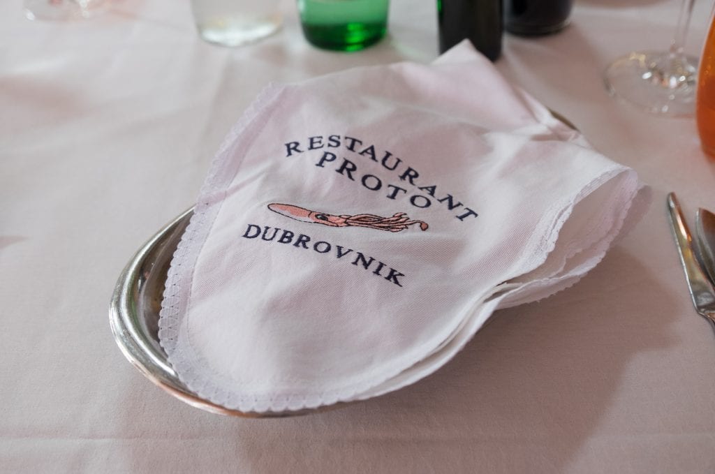 A white napkin on a silver plate. The napkin reads "Restaurant Proto Dubrovnik" with a drawing of a squid in the middle