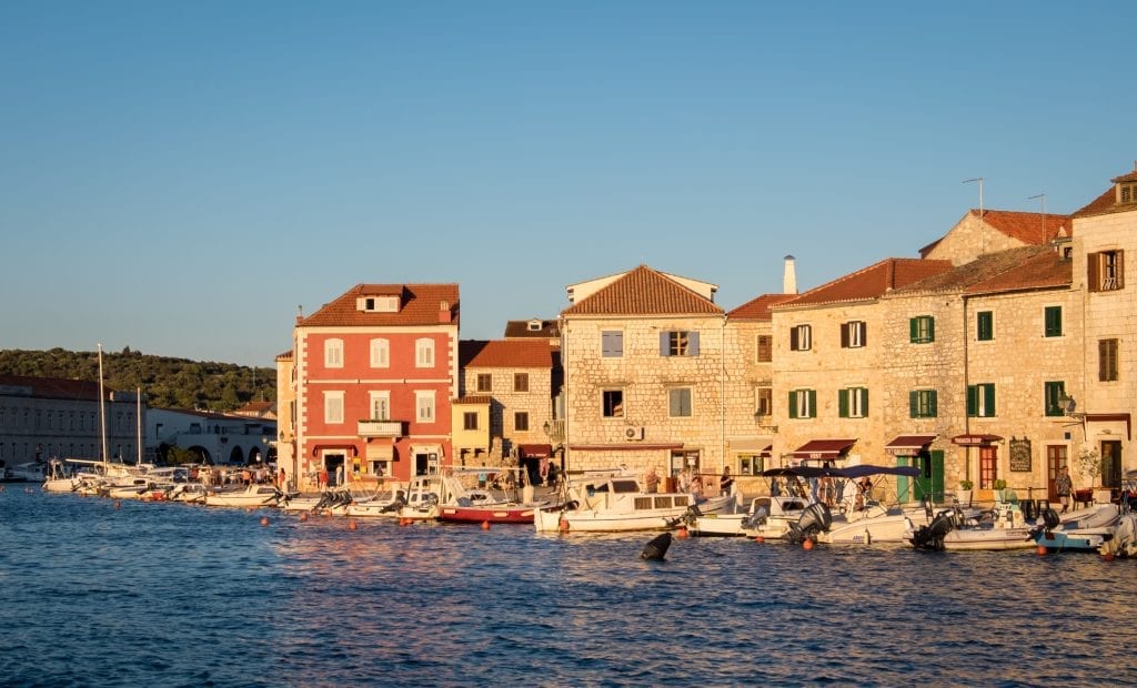 A city of sun-drenched stone buildings on the edge of the sea, lots of sailboats docked.