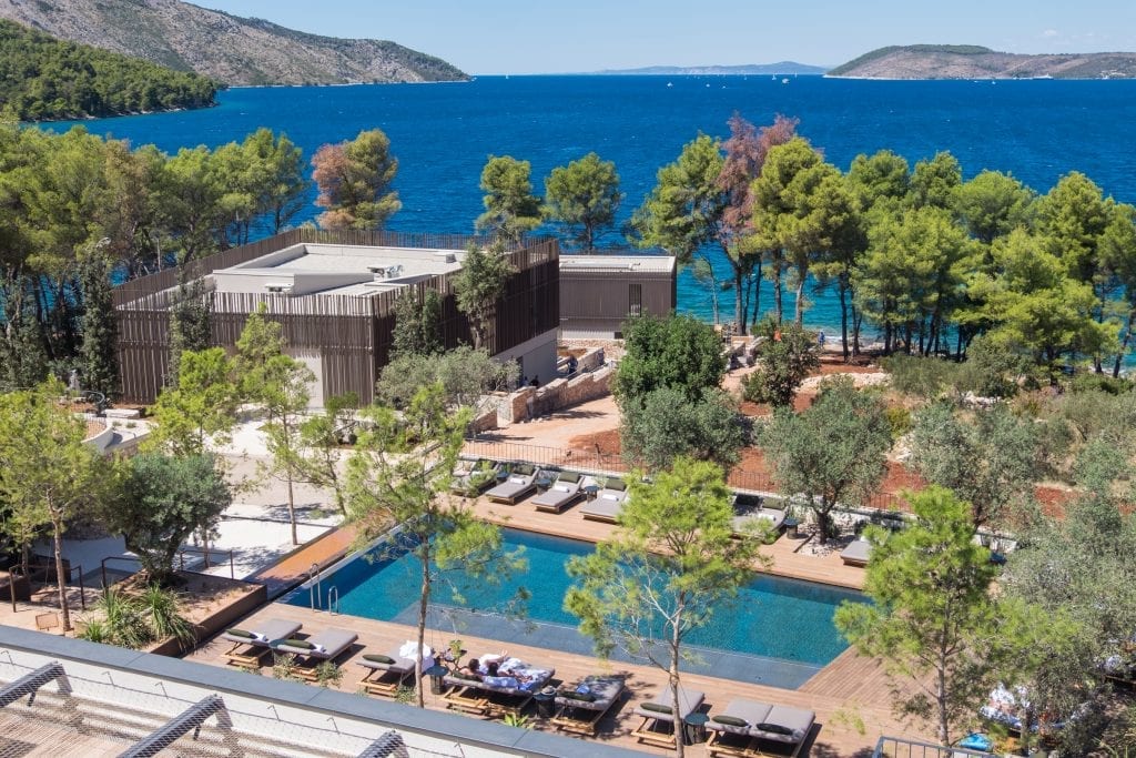 A view over the balcony at Maslina Resort Hvar. Below you see a teal rectangle pool surrounded by beach chairs and a wooden platform. There are lots of palm trees. In the background is the blue Adriatic Sea and you can see jagged coastline in the distance.