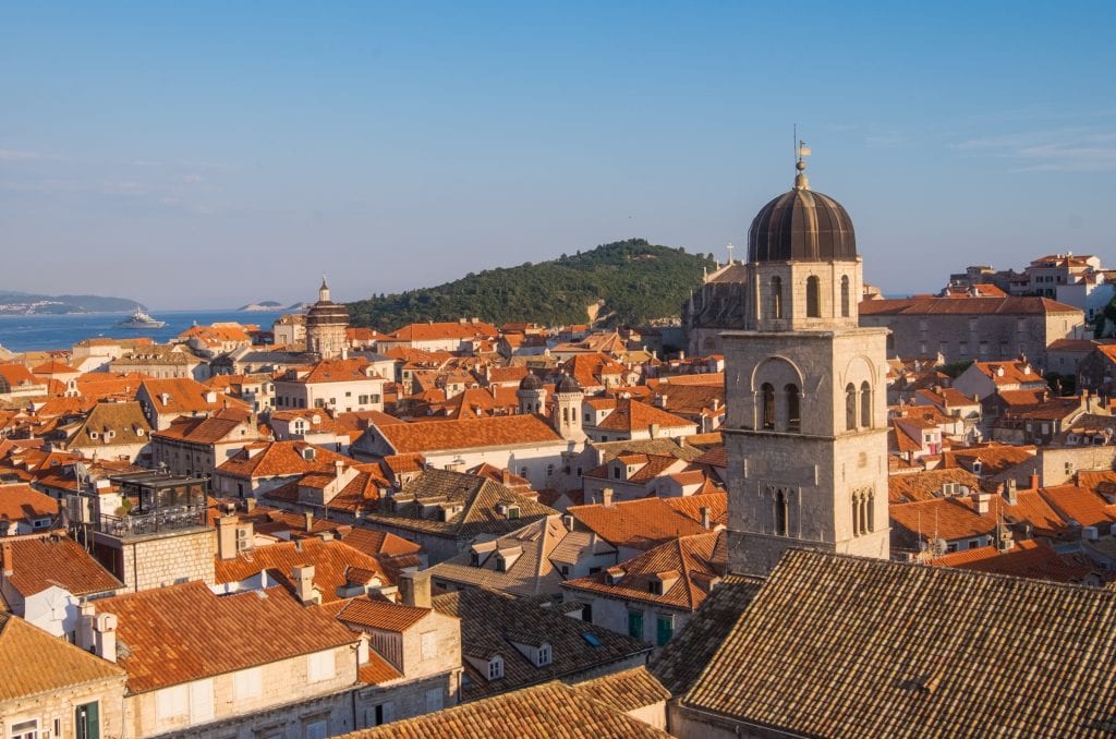 A view of the city of Dubrovnik from the walls -- lots of orange terra-cotta roofs, a church tower in the foreground, and a green island in the background, all under a blue sky.