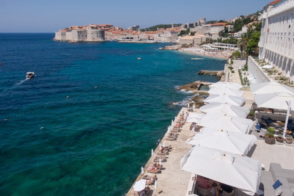 Coastline of Dubrovnik from Hotel Excelsior featuring beach chairs, white umbrellas over patio tables, and the Old Town in the background.